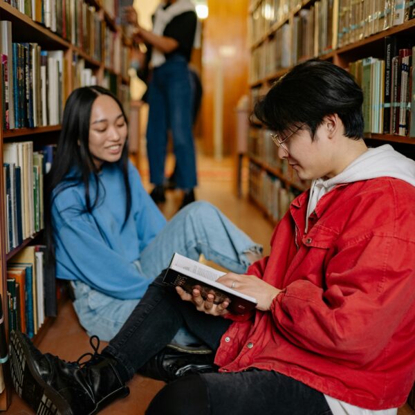 Two students sitting on a library floor reading and discussing books.